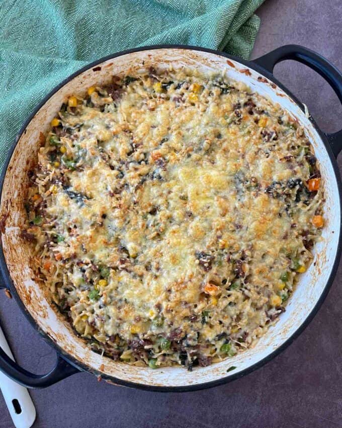 A baked casserole with melted cheese on top containing ground beef, rice, and mixed vegetables in a large round black-handled baking dish.