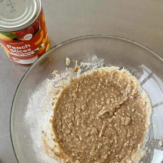 Peach Cobbler A glass bowl with a mixed batter sits next to an open can of peach slices in light syrup on a stainless steel surface.