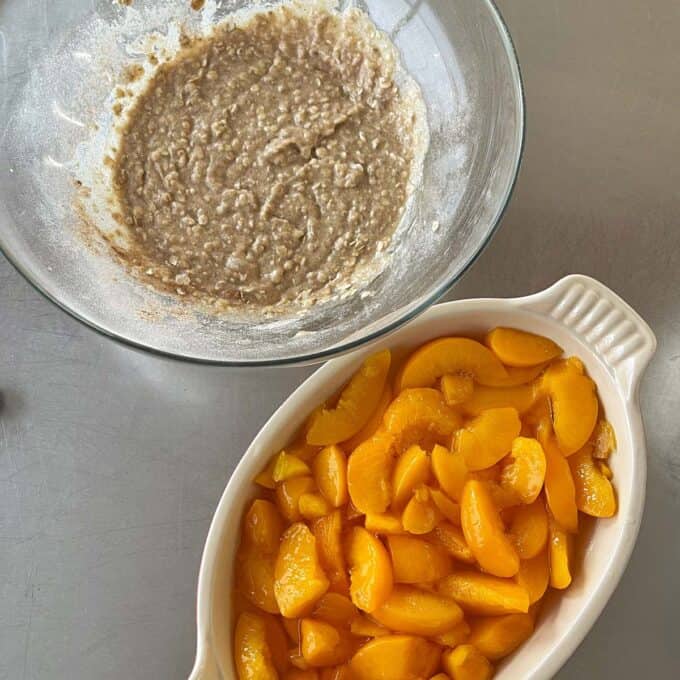 Peach Cobbler A glass bowl with oatmeal batter next to a white dish filled with sliced canned peaches on a gray countertop.