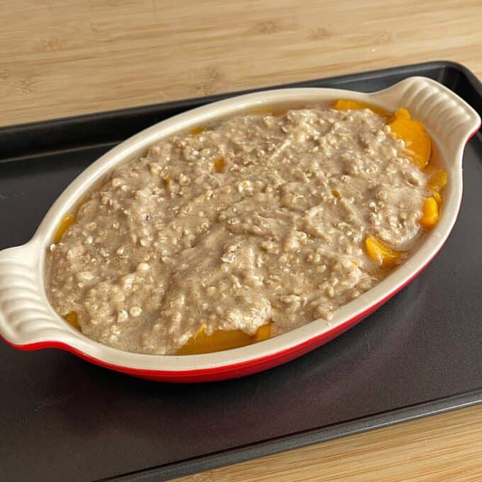 Peach Cobbler An oval baking dish with sliced peaches covered in an oatmeal batter sits on a black baking tray on a wooden surface.