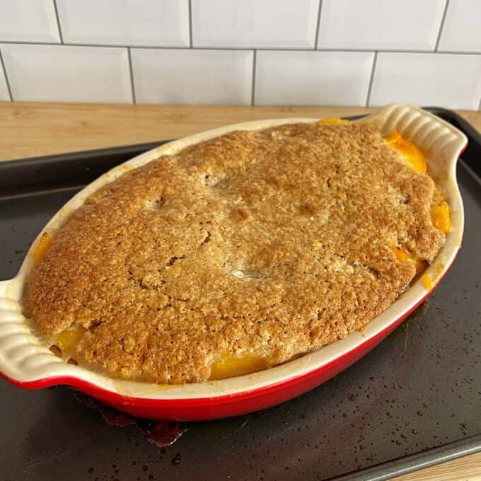 Peach Cobbler A baked peach cobbler in a red oval dish on a black baking tray, with a golden-brown crust and some filling visible at the edges. White tile backsplash in the background.