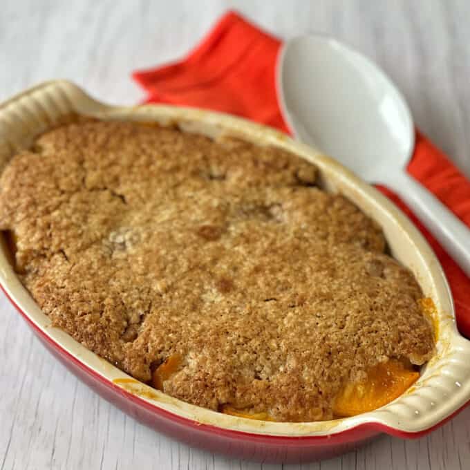 Peach Cobbler A baked peach cobbler with a golden-brown crust in an oval dish, placed next to a white serving spoon and a red cloth napkin on a light surface.