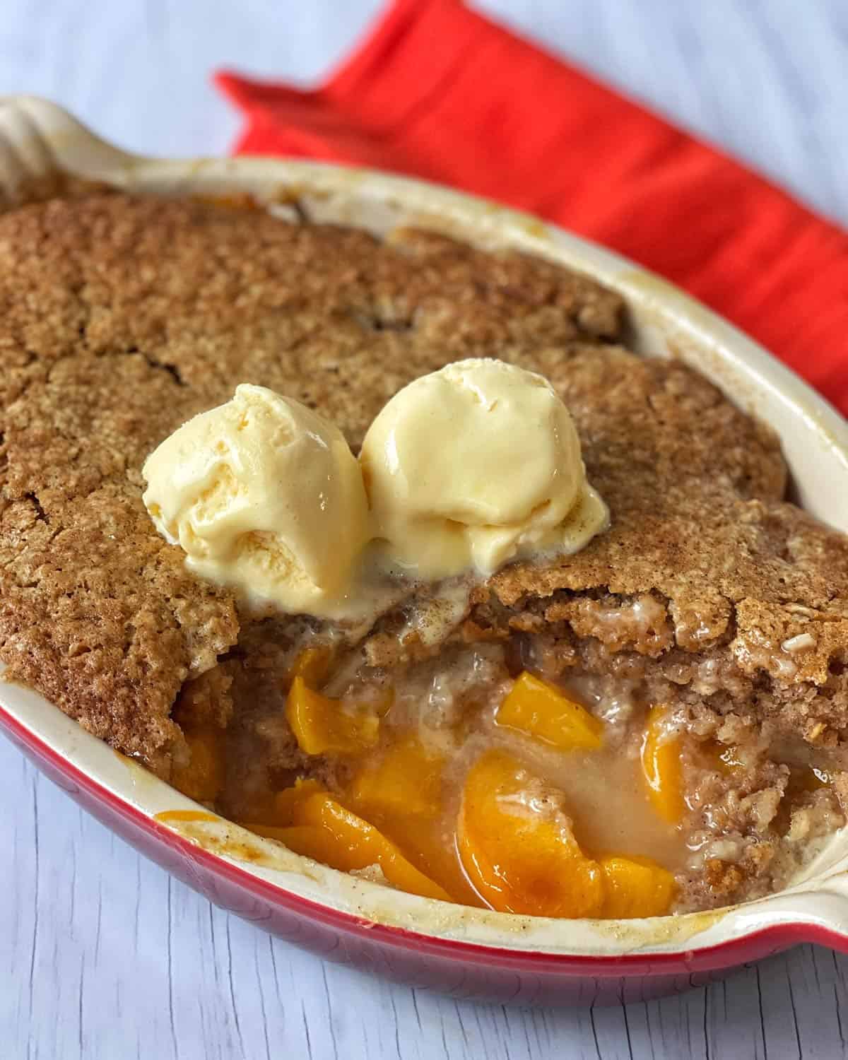 Peach Cobbler A baked peach cobbler in a dish, topped with two scoops of vanilla ice cream; a red napkin is placed beside the dish.