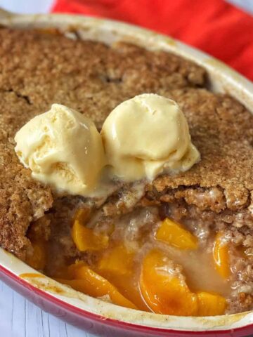 A peach cobbler topped with two scoops of vanilla ice cream, with a portion scooped out to reveal the filling.