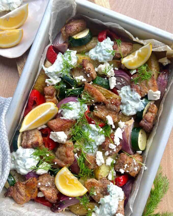 Pork Souvlaki Tray Bake A tray of roasted vegetables, chicken pieces, lemon wedges, and tzatziki sauce, garnished with fresh dill and crumbled feta cheese.