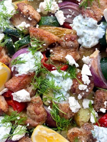 Tray of roasted vegetables, grilled meat pieces, lemon wedges, fresh dill, feta cheese crumbles, and dollops of creamy white sauce.