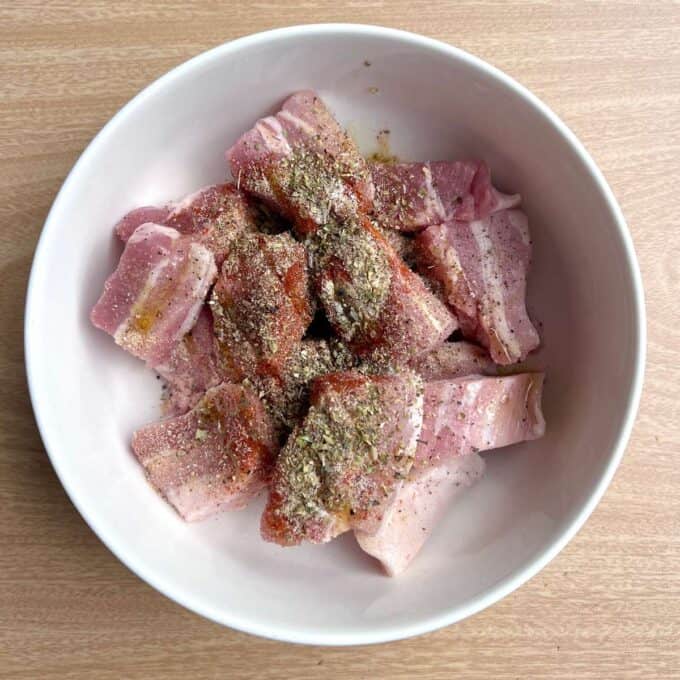 Pork Souvlaki Tray Bake Raw pork belly slices in a white bowl, sprinkled with various dried herbs, spices, salt, and pepper, on a wooden surface.