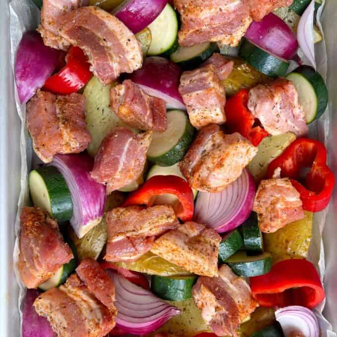 Pork Souvlaki Tray Bake Chopped bacon, red onion, zucchini, red bell pepper, and potatoes, seasoned and arranged on a baking tray, ready for roasting.