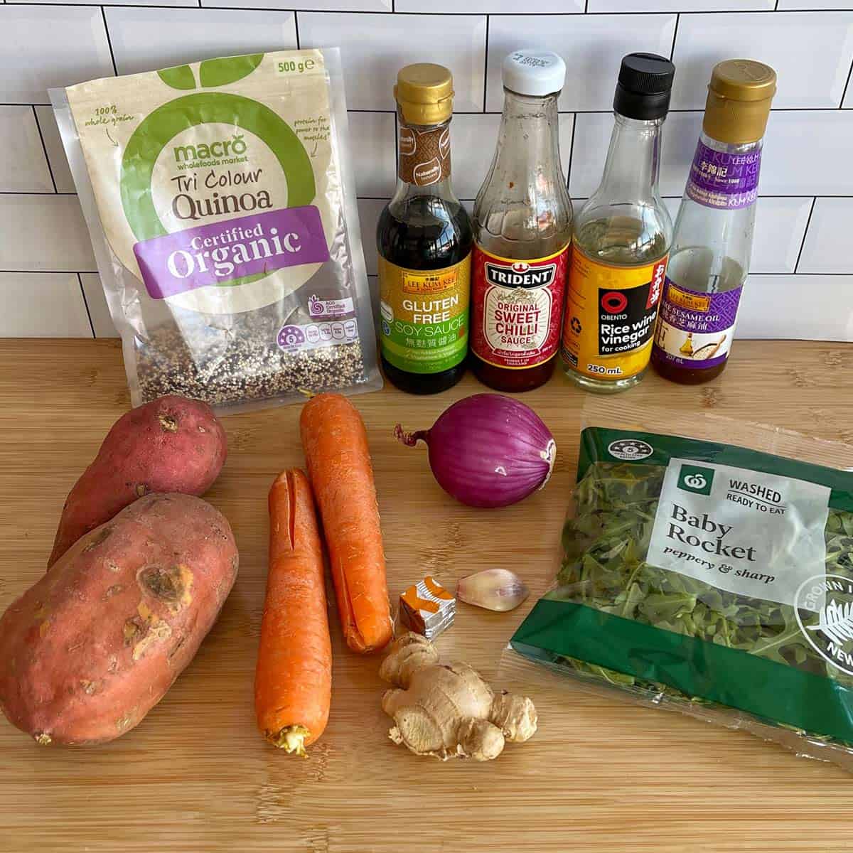 Quinoa, Kumara and Carrot Salad A selection of ingredients on a wooden surface, including sweet potatoes, carrots, red onion, garlic, ginger, salad greens, quinoa, and various sauces and vinegar bottles.