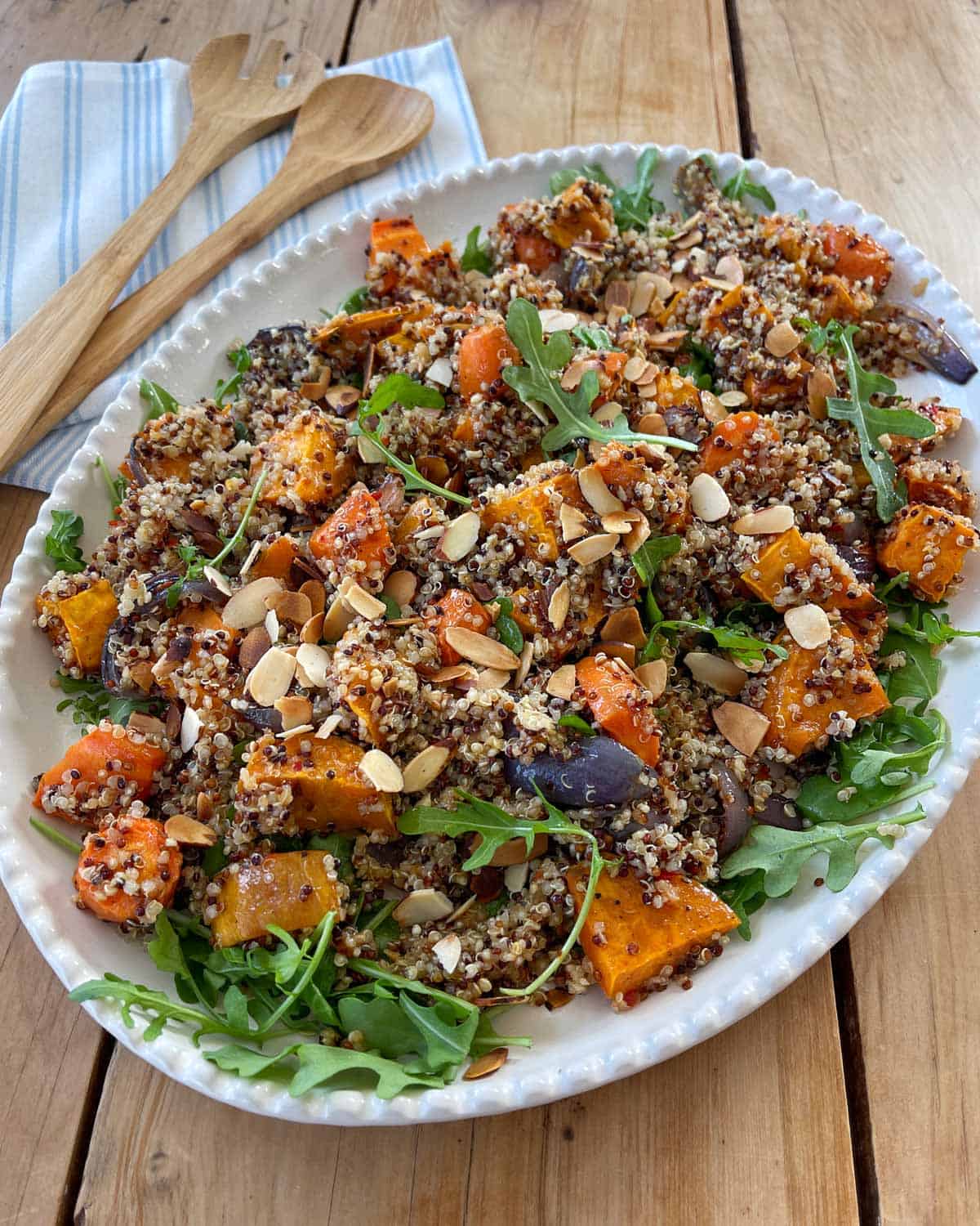 Quinoa, Kumara and Carrot Salad A platter of quinoa salad with roasted butternut squash, red onions, arugula, and sliced almonds on a wooden table with wooden serving utensils.