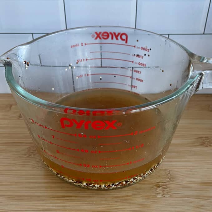 Quinoa, Kumara and Carrot Salad A glass Pyrex measuring cup partially filled with brown liquid and some seasoning particles, placed on a wooden surface with a tiled wall background.