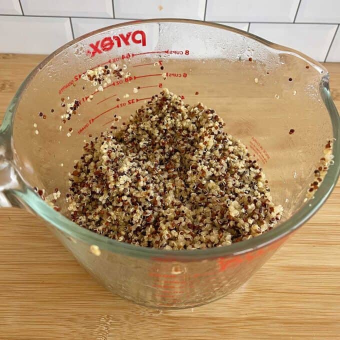 Quinoa, Kumara and Carrot Salad Cooked tri-color quinoa in a clear glass Pyrex measuring cup on a wooden surface, with a white tile backsplash in the background.