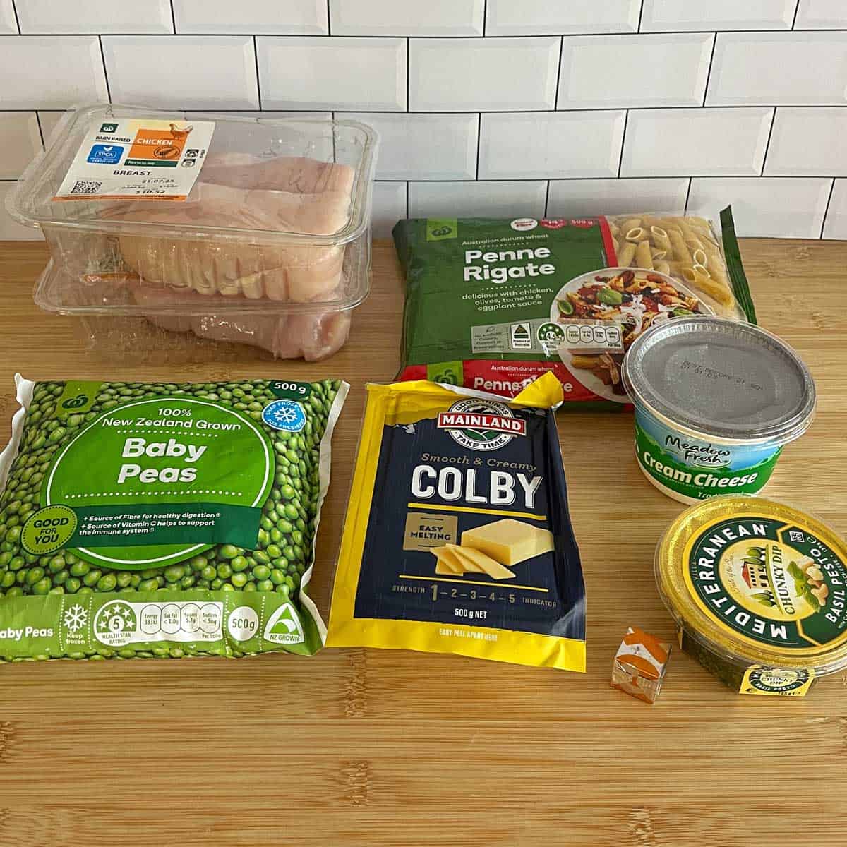 A selection of food items on a wooden surface, including raw fish fillets, penne pasta, baby peas, Colby cheese, cream cheese, wedge cheese, and a tub of spreadable butter.
