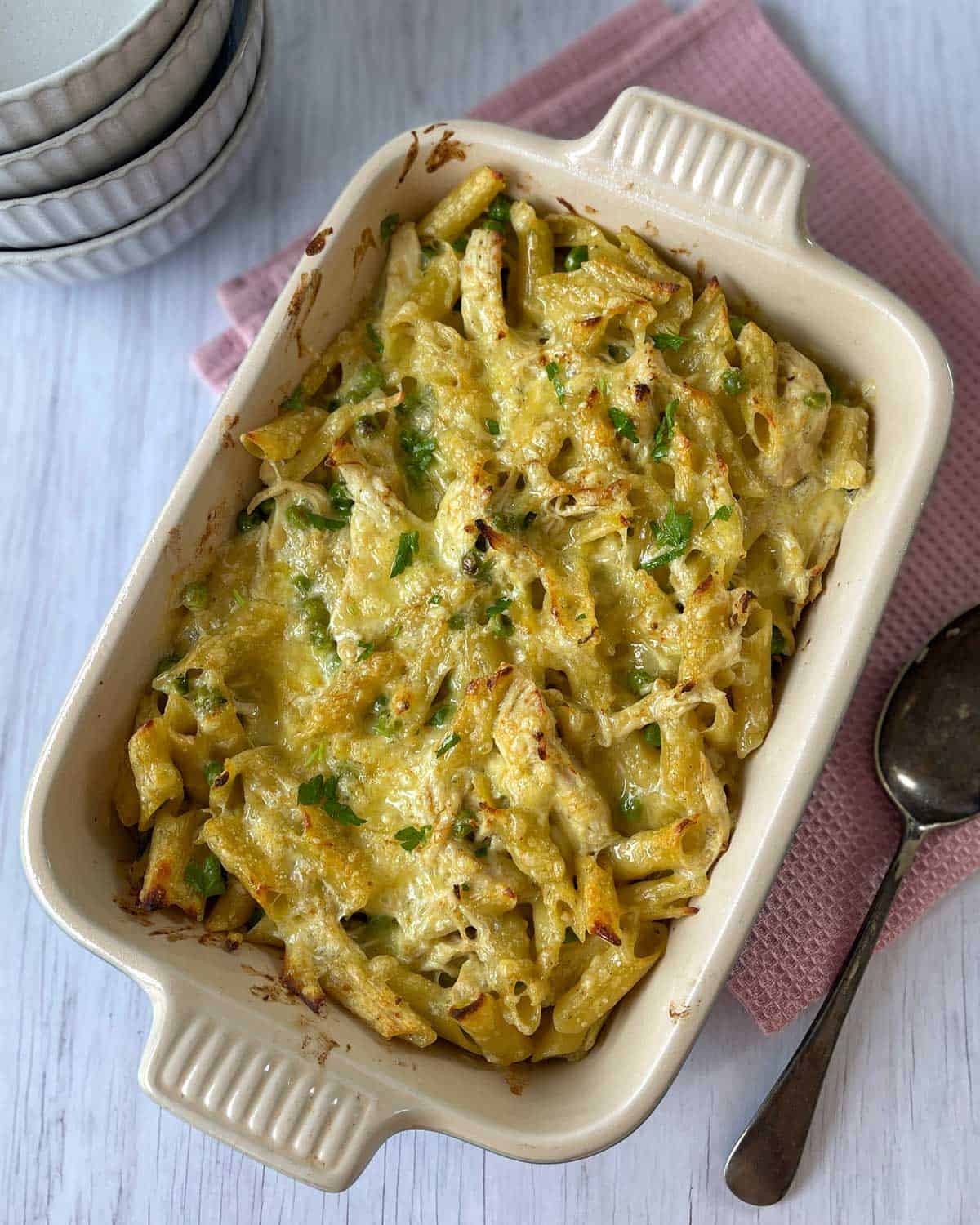 Rectangular baking dish with baked pasta topped with melted cheese and herbs, placed on a pink cloth next to stacked bowls and two spoons.