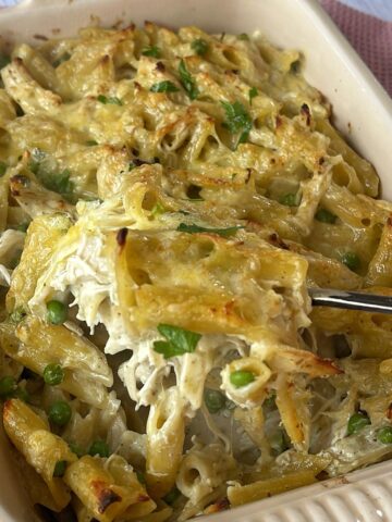 Baked pasta casserole with penne, green peas, shredded chicken, and melted cheese being served from a rectangular dish.