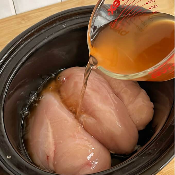 Raw chicken breasts in a slow cooker with broth being poured from a measuring cup.