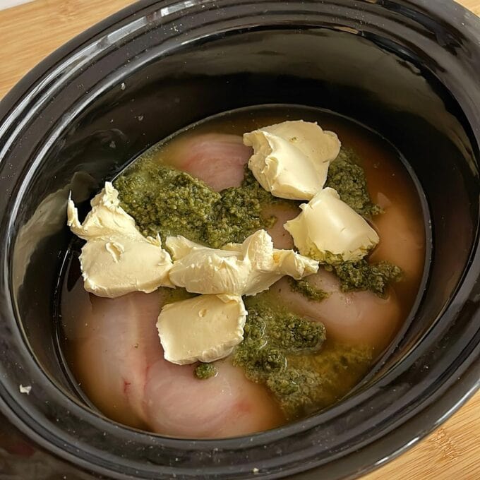 Raw chicken breasts in a slow cooker with dollops of cream cheese, pesto sauce, and some liquid at the bottom.