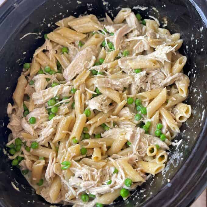 Cooked penne pasta mixed with shredded chicken, green peas, and a creamy white sauce in a black slow cooker.
