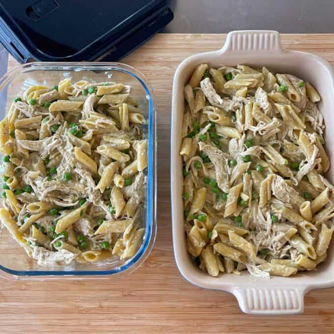 Two containers of baked penne pasta mixed with shredded chicken, green peas, and creamy sauce are placed on a wooden surface.