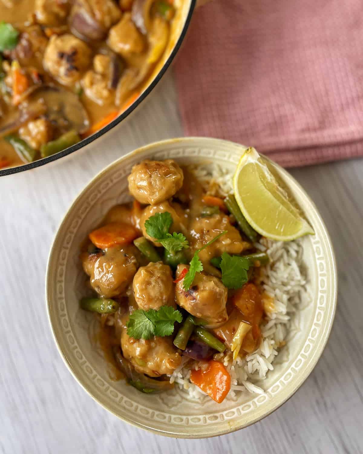 Satay Sausage Rice Bowl A bowl of rice topped with vegetable curry, garnished with cilantro and a lime wedge, with extra curry in a pan nearby. A pink napkin is placed to the side.