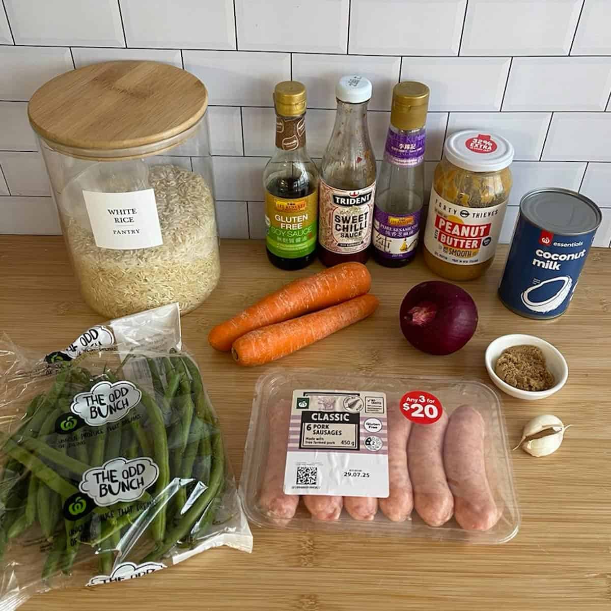 Satay Sausage Rice Bowl Ingredients for a meal arranged on a wooden surface, including rice, sauces, peanut butter, coconut milk, green beans, carrots, onion, sausages, brown sugar, and garlic.