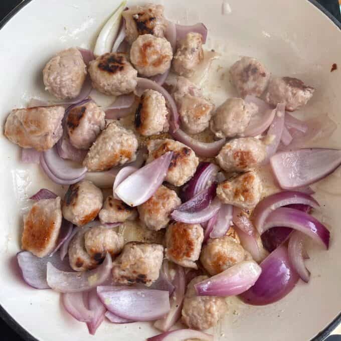 Satay Sausage Rice Bowl Meatballs and sliced red onions cooking in a white skillet, with some pieces browned and onions slightly softened.