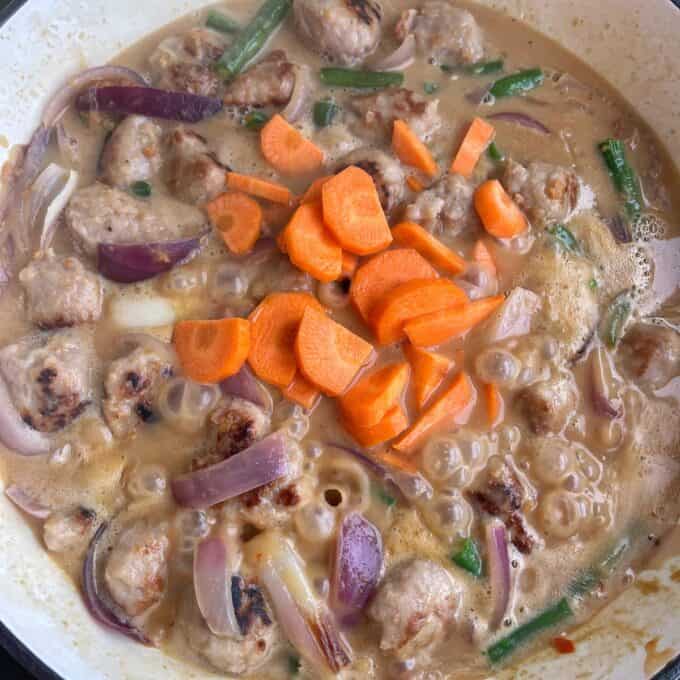 Satay Sausage Rice Bowl Meatballs simmering in a creamy sauce with sliced carrots, red onions, and green beans in a pan.