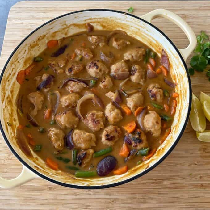 Satay Sausage Rice Bowl A cream-colored pot filled with a brown curry containing meatballs, sliced red onions, carrots, and green beans on a wooden surface. Lemon wedges and cilantro are nearby.