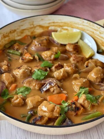 A pot of vegetable and tofu curry in a creamy sauce, garnished with cilantro and lime wedges, with a serving spoon in the dish.