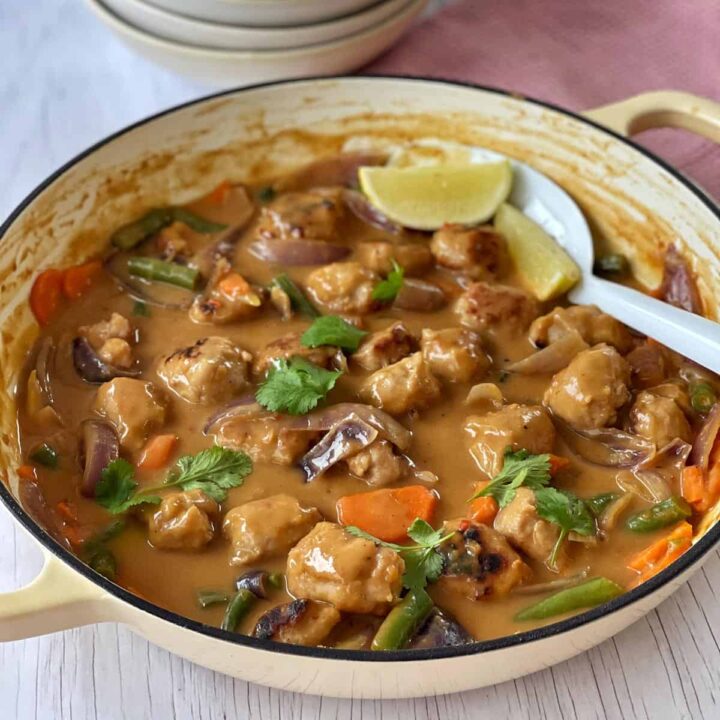 A pot of vegetable and tofu curry in a creamy sauce, garnished with cilantro and lime wedges, with a serving spoon in the dish.