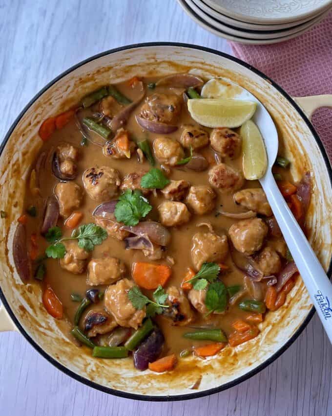 Satay Sausage Rice Bowl A pot of creamy curry with chicken pieces, carrots, green beans, red onions, garnished with cilantro and lemon wedges, with a spoon resting on the side.