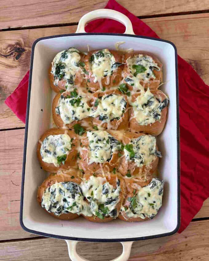 Creamy Spinach Cob Sliders A white baking dish with nine bread rolls topped with melted cheese, spinach, and green peppers, placed on a wooden surface with a red cloth underneath.