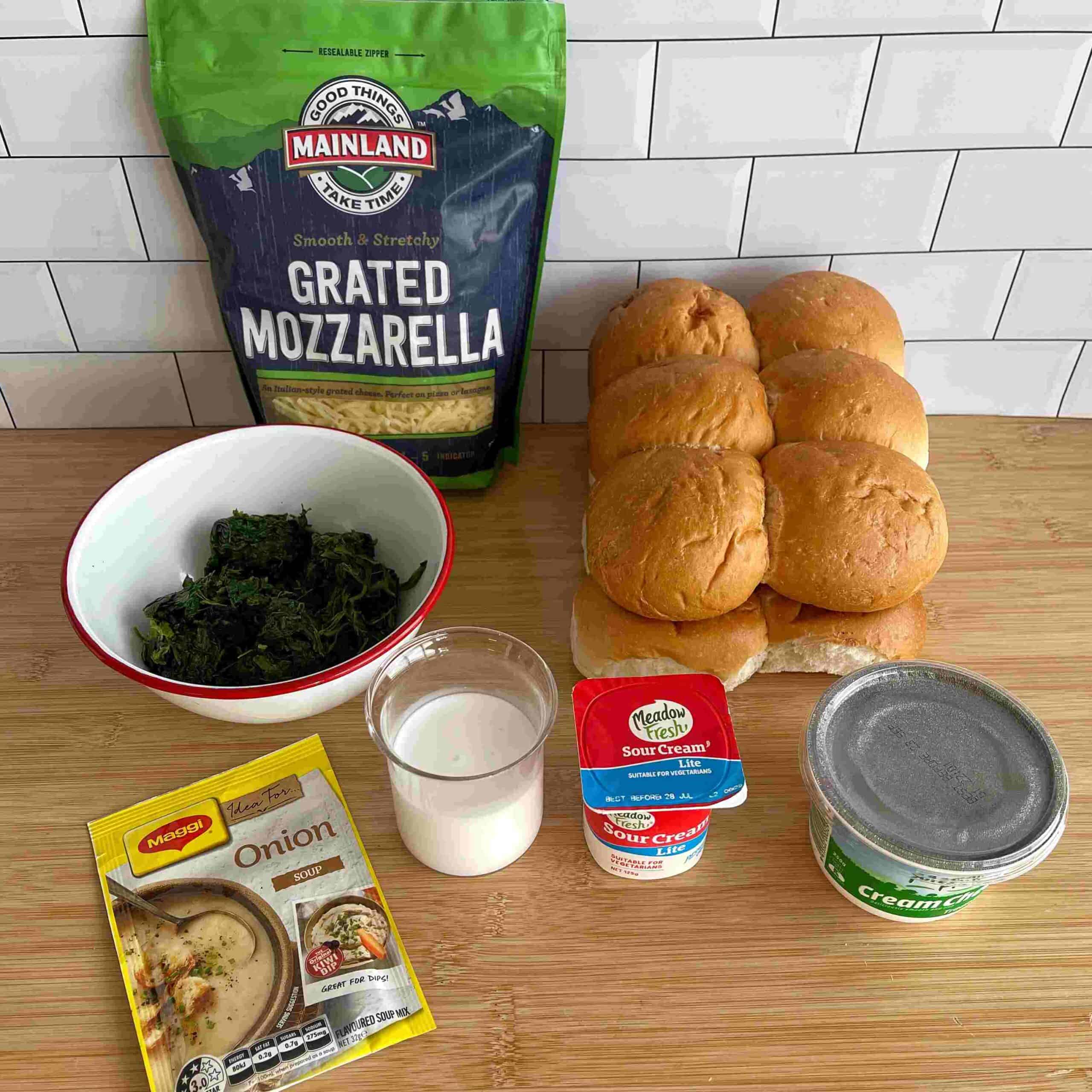 Creamy Spinach Cob Sliders Ingredients for a recipe on a wooden surface: grated mozzarella, six bread rolls, frozen spinach, onion soup mix, milk, sour cream, and a can of cream cheese.