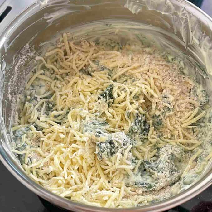 Creamy Spinach Cob Sliders A metal mixing bowl containing uncooked shredded potatoes, spinach, creamy sauce, and seasonings, being prepared for a recipe.