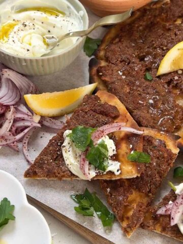 A tray with slices of meat-topped flatbread garnished with herbs and onions, served with lemon wedges, sliced red onions, and a bowl of yogurt with a spoon.
