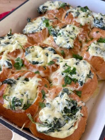 A baking dish filled with Creamy Spinach Cob Sliders-bread rolls topped with melted cheese, spinach, and herbs-sits on a wooden surface with a red cloth nearby.