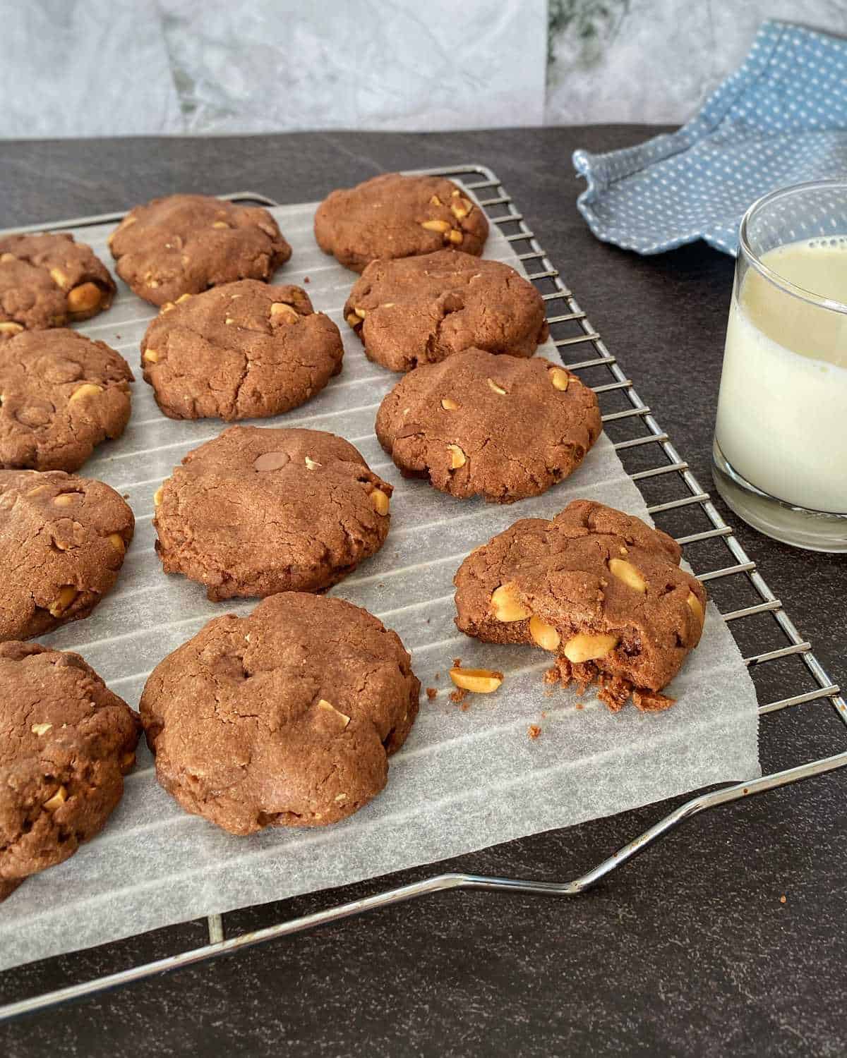 A batch of chocolate chip cookies with nuts on a cooling rack lined with parchment paper, next to a glass of milk on a dark countertop.
