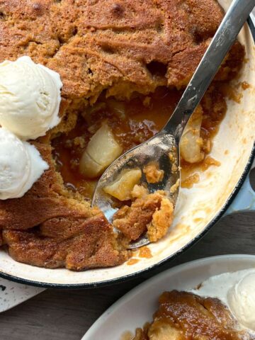 A pan of baked cobbler with a golden-brown crust, a serving spoon, and two scoops of vanilla ice cream. A portion is served on a plate with more ice cream.