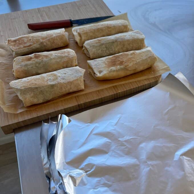 Freezer-Friendly Breakfast Burritos Six wrapped burritos on parchment paper on a wooden cutting board, with a knife nearby and a sheet of aluminum foil in the foreground.