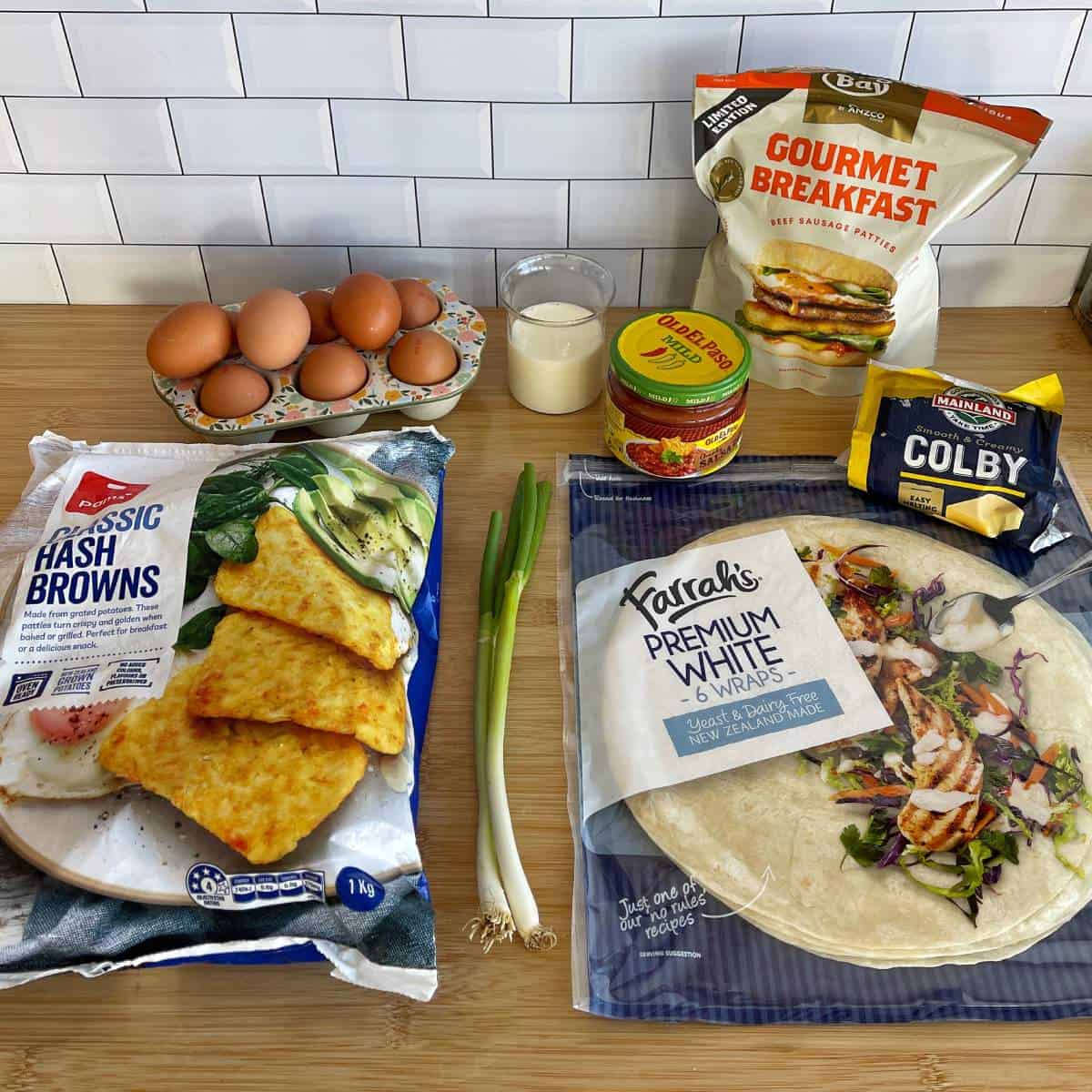 Freezer-Friendly Breakfast Burritos A selection of breakfast ingredients on a countertop, including eggs, milk, hash browns, wraps, green onions, shredded cheese, and a bag of breakfast sausage patties.