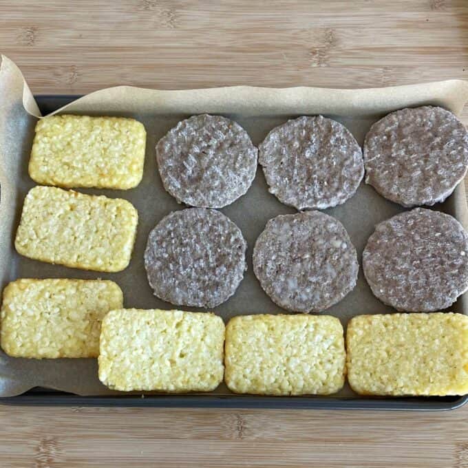 Freezer-Friendly Breakfast Burritos A baking tray lined with parchment paper holds six rectangular frozen hash browns and six round frozen sausage patties arranged in rows.