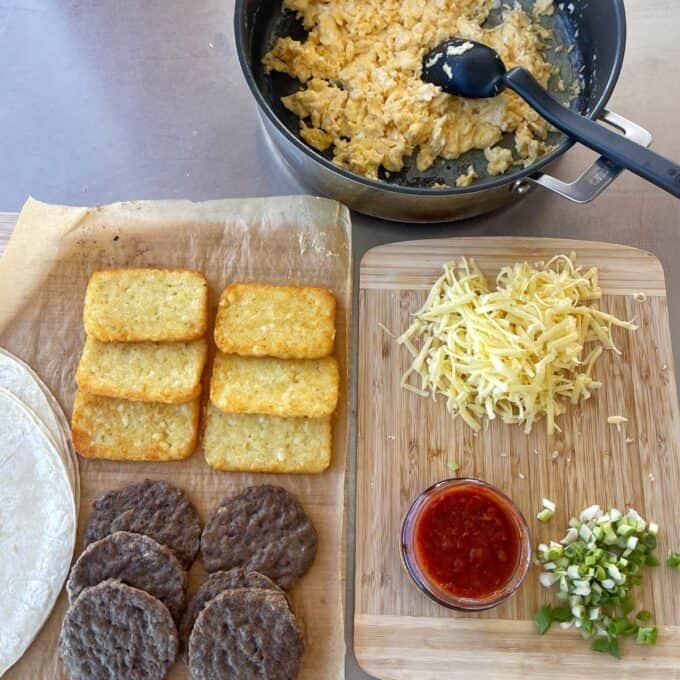 Freezer-Friendly Breakfast Burritos A cutting board with shredded cheese, salsa, and chopped green onions next to cooked hash browns, sausage patties, tortillas, and a pan of scrambled eggs.
