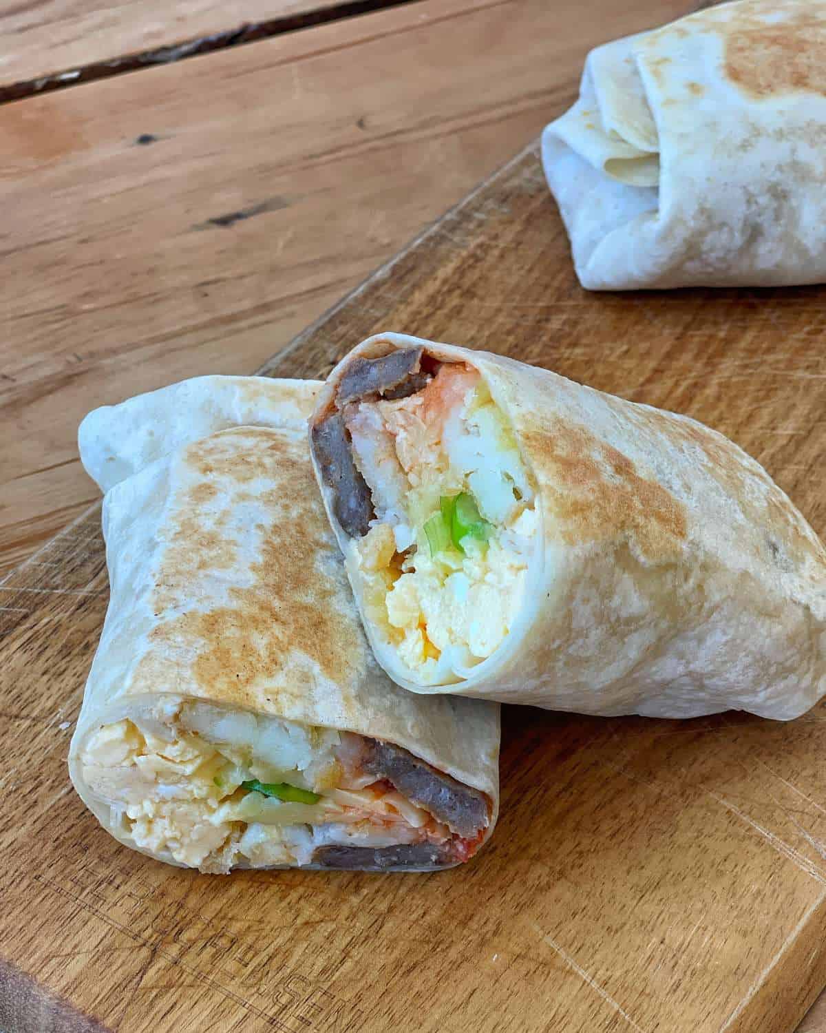 Two breakfast burritos cut in half, showing fillings of eggs, beans, potatoes, and vegetables, placed on a wooden cutting board.