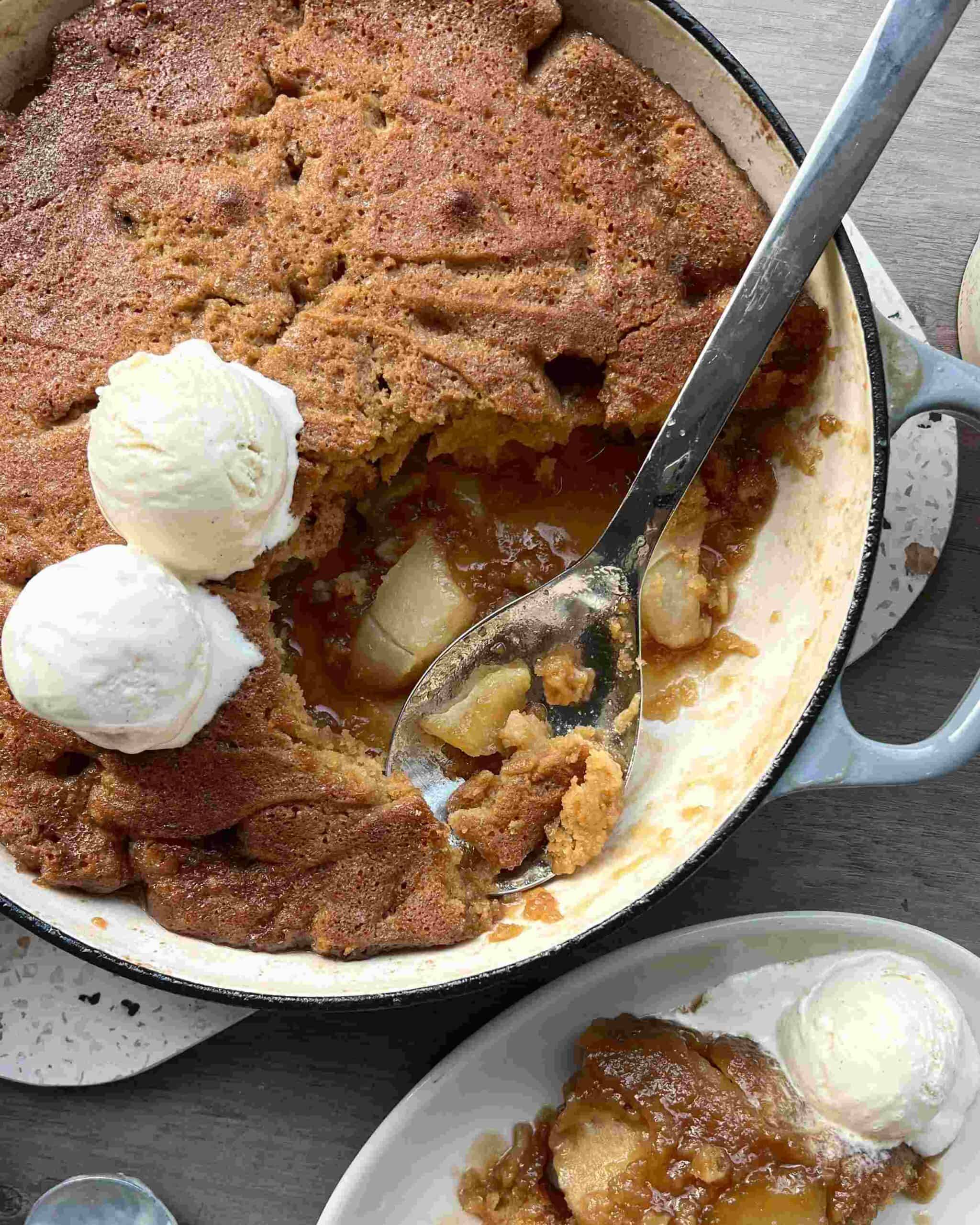 A baked cobbler with a golden crust and fruit filling is served in a round dish with two scoops of vanilla ice cream; a portion is plated alongside.