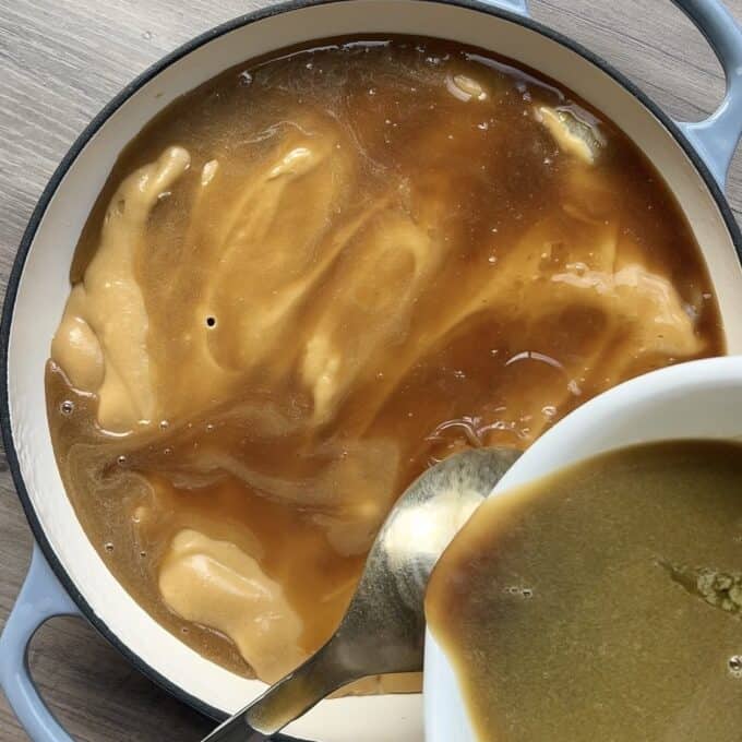 Caramel Apple Sponge Pudding A spoon pours brown gravy from a white bowl into a pot filled with more gravy on a wooden surface.