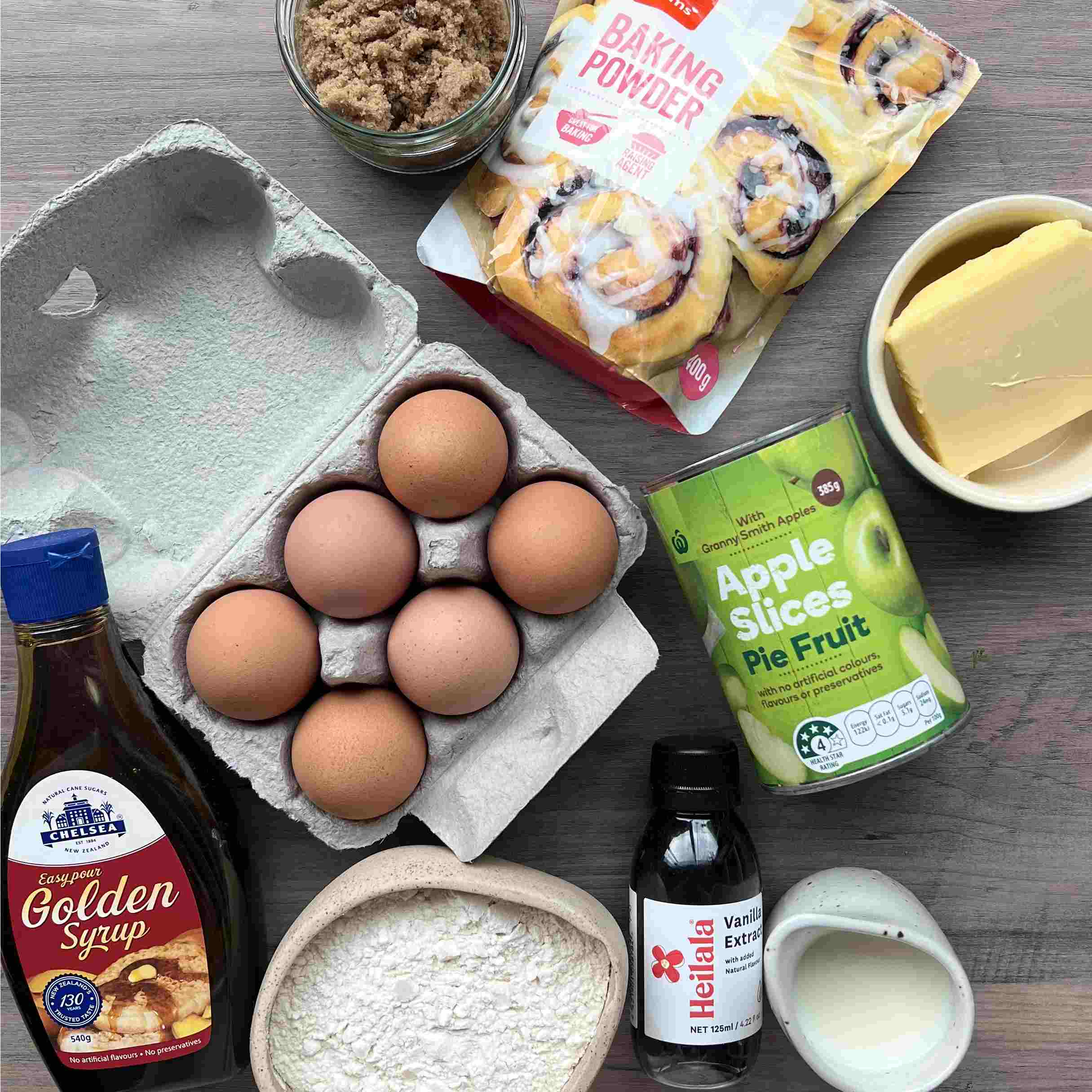 Caramel Apple Sponge Pudding Baking ingredients including eggs, flour, butter, brown sugar, baking powder, golden syrup, vanilla extract, milk, a can of apple slices, and a packet of cinnamon rolls arranged on a table.