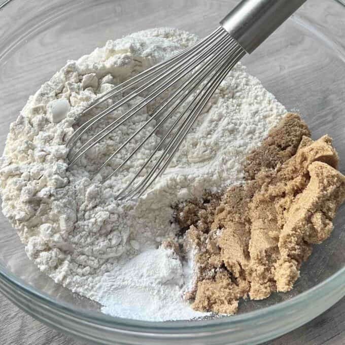 Caramel Apple Sponge Pudding A metal whisk rests in a glass bowl containing flour, brown sugar, and baking powder, ready to be mixed.