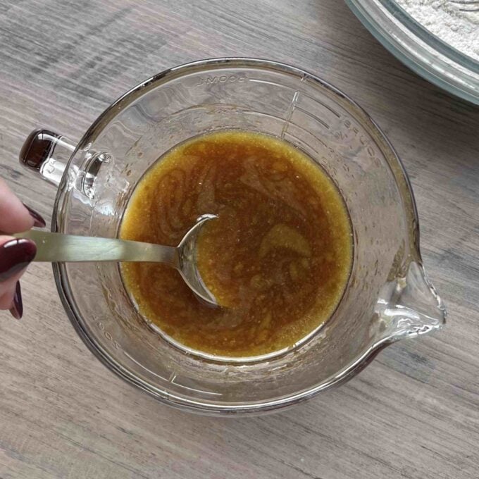 Caramel Apple Sponge Pudding A hand stirs a brown liquid mixture in a glass measuring cup with a metal spoon on a wooden surface.