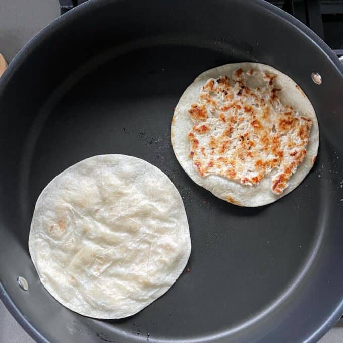 Homemade McChicken Tacos Two corn tortillas are being heated in a black nonstick pan, with one tortilla toasted and browned while the other is still pale.