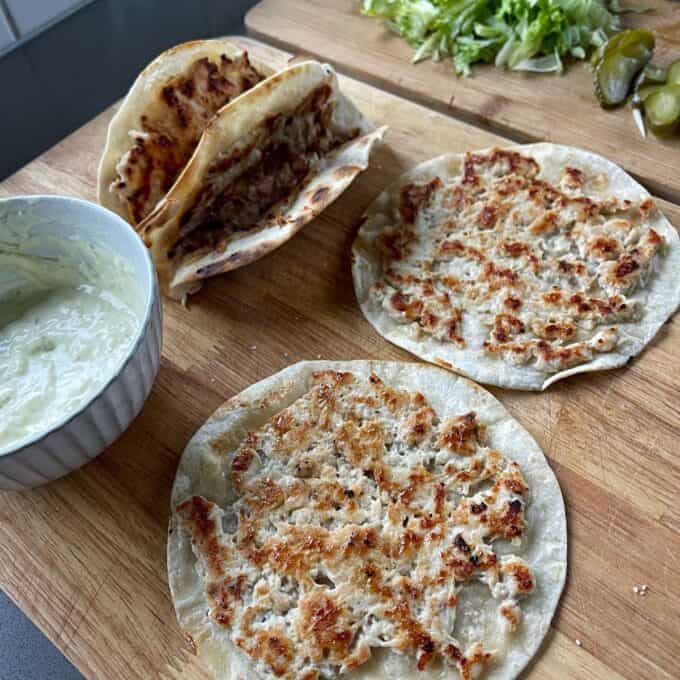Homemade McChicken Tacos Two flatbreads with grilled minced meat next to folded wraps, a bowl of white sauce, sliced lettuce, and pickles on a wooden board.